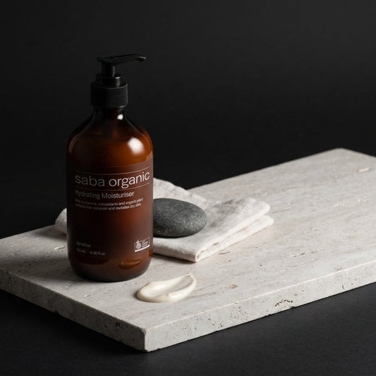 Amber glass bottle of Saba Organic Hydrating Moisturiser on pale stone with a swirl of cream, softly lit against a dark background.